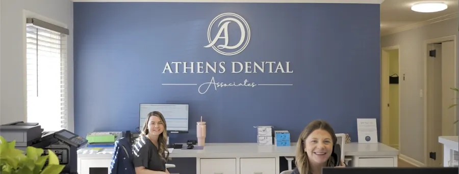 Two Athens Dental Associates team members at the front desk, with the practice's navy logo wall behind them.